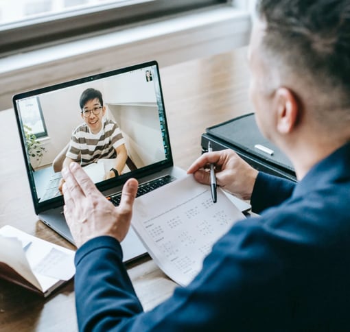 a teacher at his desk, holding a calendar and a pen in his hand while talking with a student during a video call on his laptop